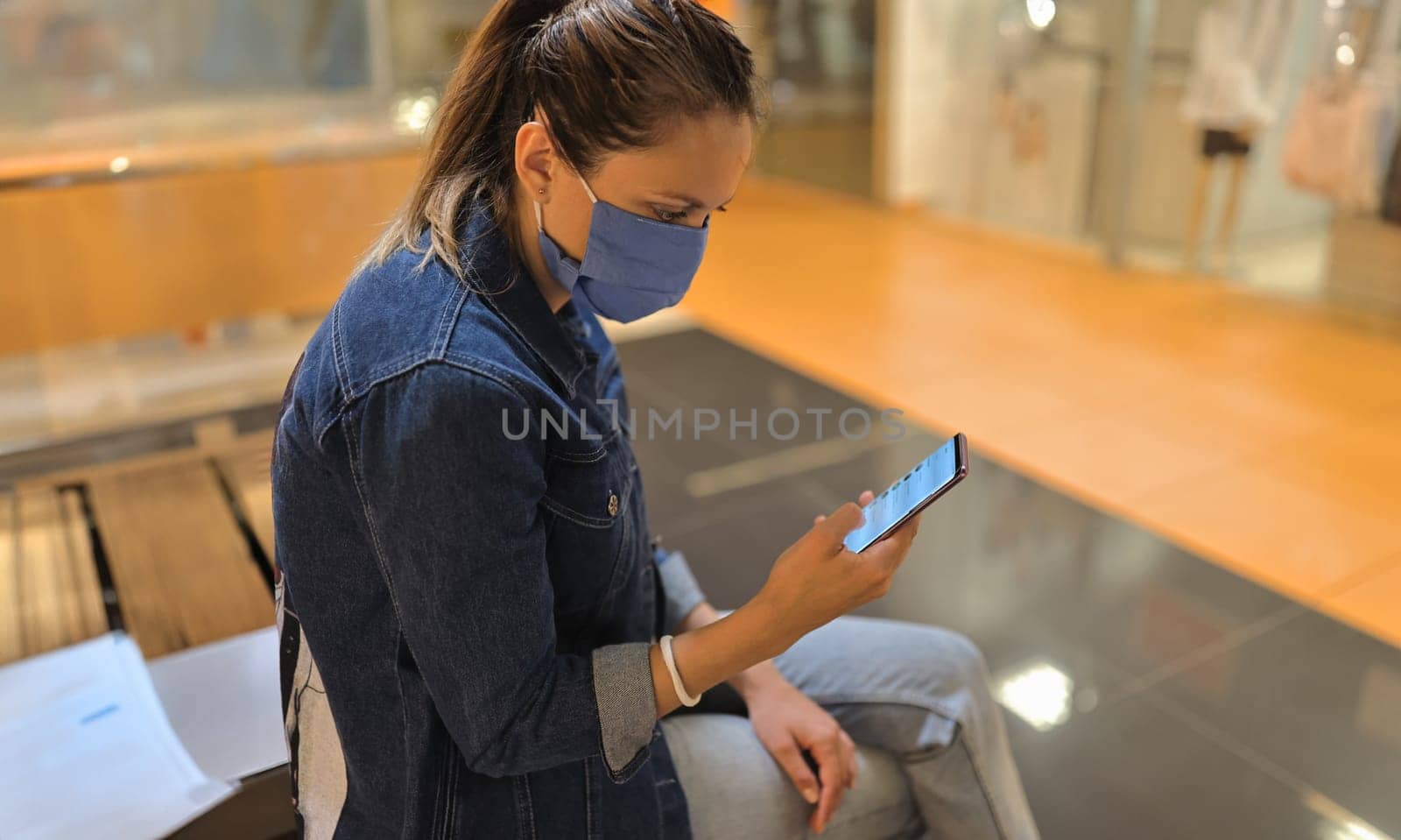 Woman in protective mask sit on bench in shopping center and look at phone. Empty hall in trading floor, showcases with manikin.