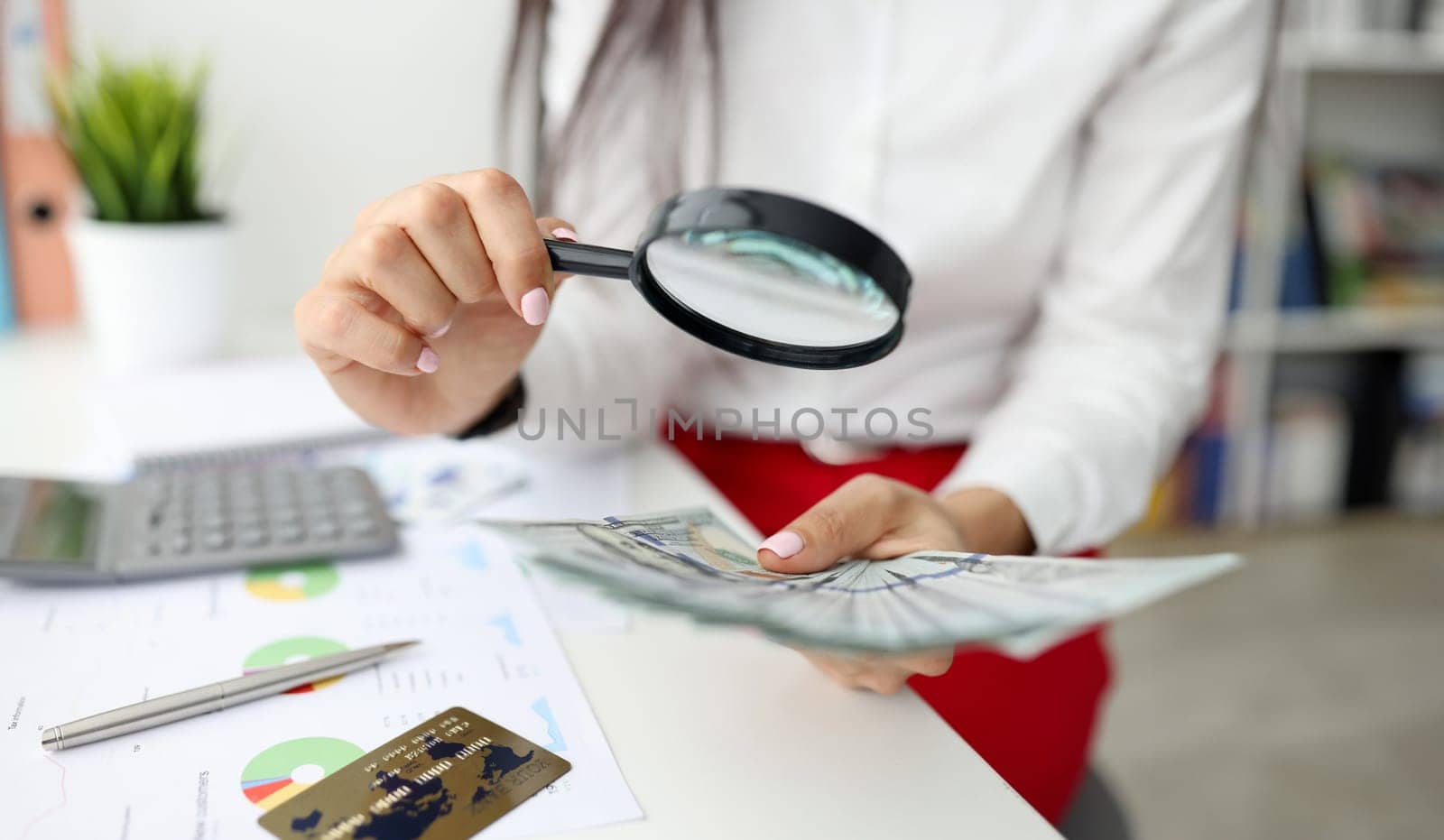 Woman check currency for authenticity. Female hand hold magnifying glass and lot of money. On white table lies gray calculator, metal pen, bank card and document with diagram.