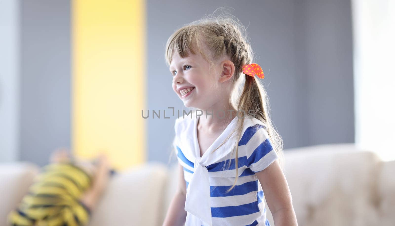 Happy girl in striped T-shirt and with pigtails laugh. In background child indulge on couch.