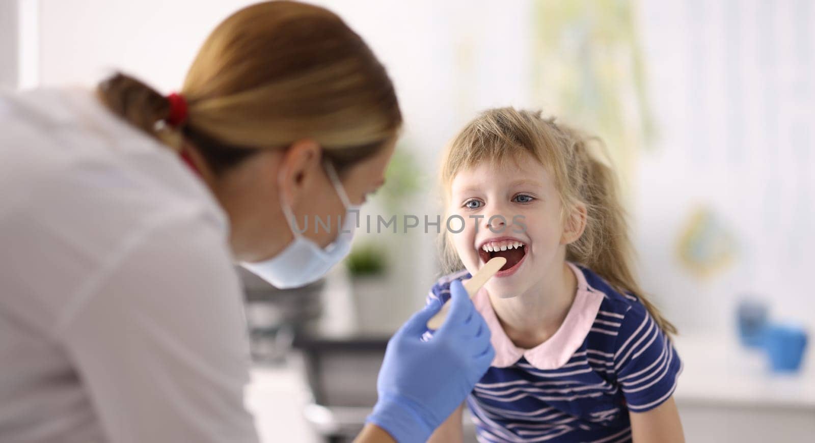 Woman doctor pediatrician in protective medical mask and rubber gloves examines throat of little girl wooden spatula portrait. Examination and treatment of children in pandemic covid 19 concept.