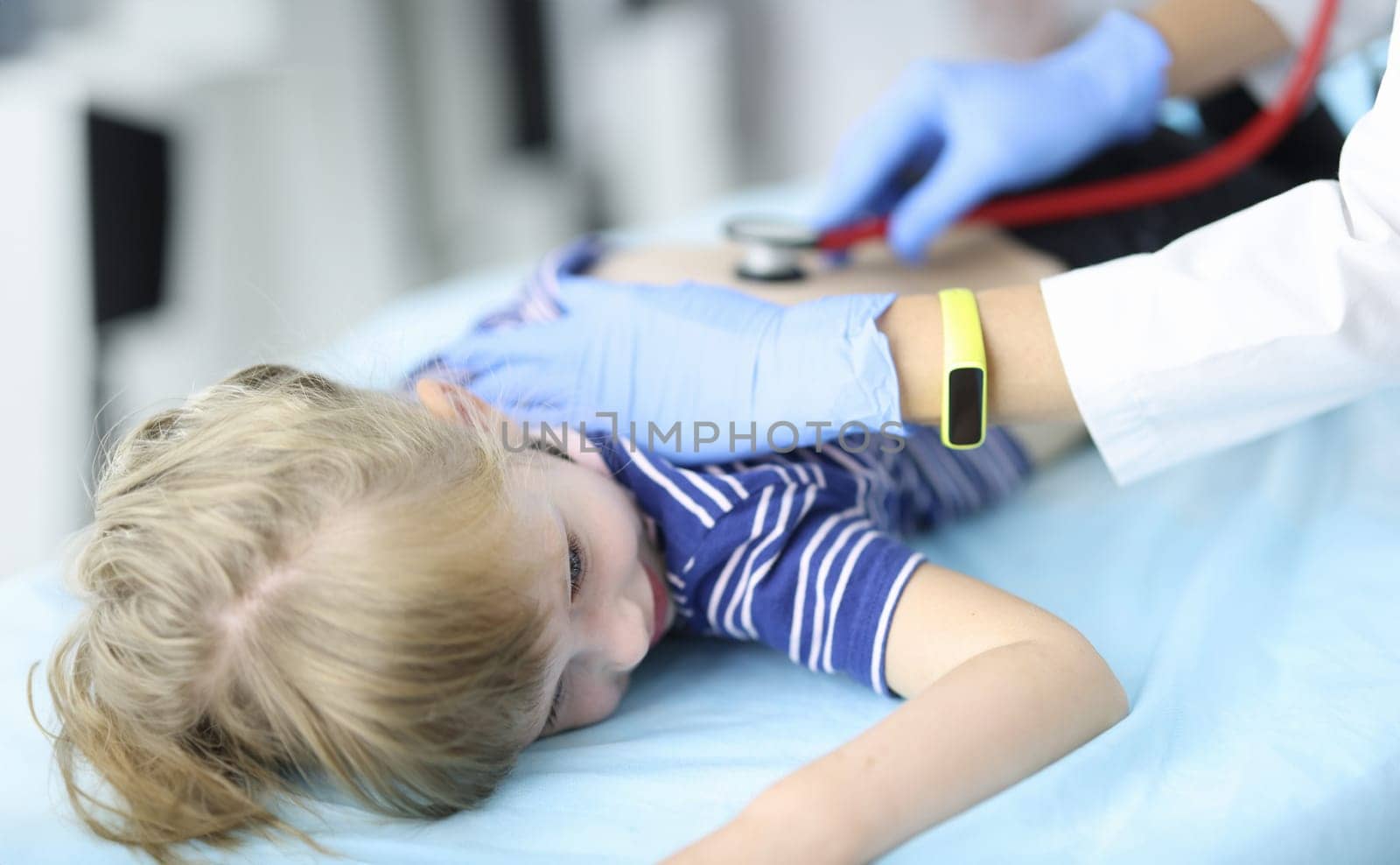 Pediatrician doctor in rubber glove listens to lungs of child lying on stomach on couch in clinic portrait by kuprevich