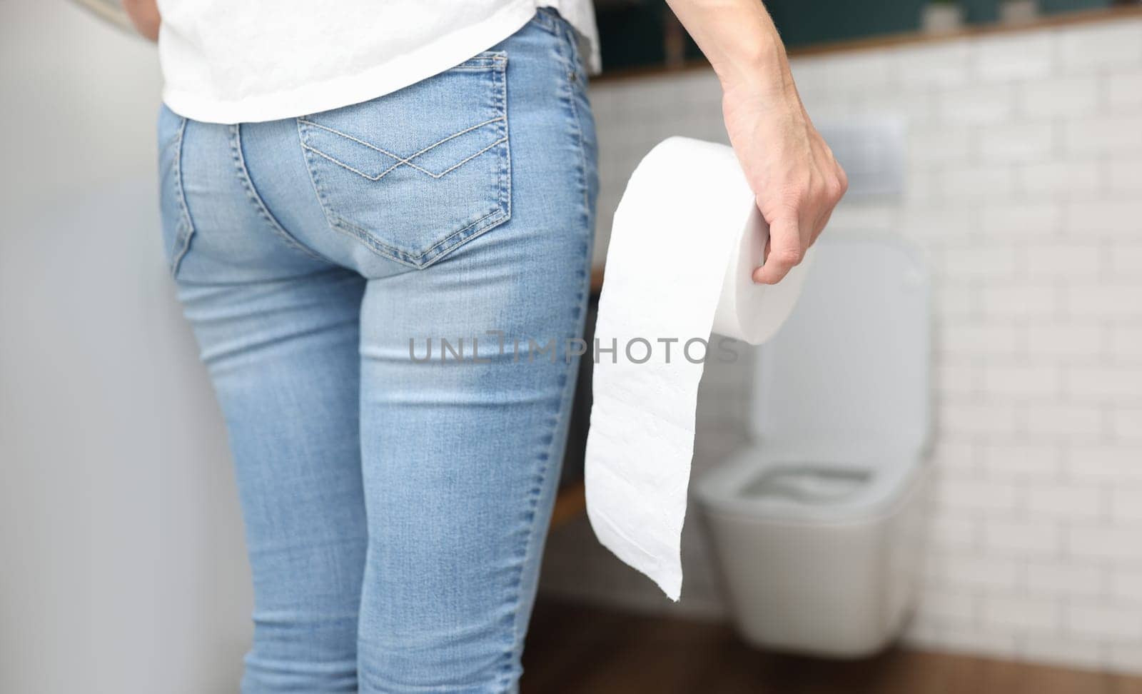 Woman in blue jeans stands with back and holds roll of toilet paper in her hand in front of toilet close-up. Frequent urination urology concept.
