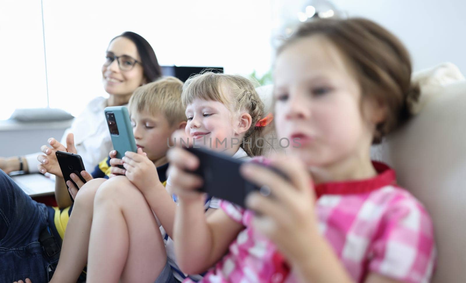 Three children sit side by side on couch and play on smartphones. Woman work at laptop next to children.