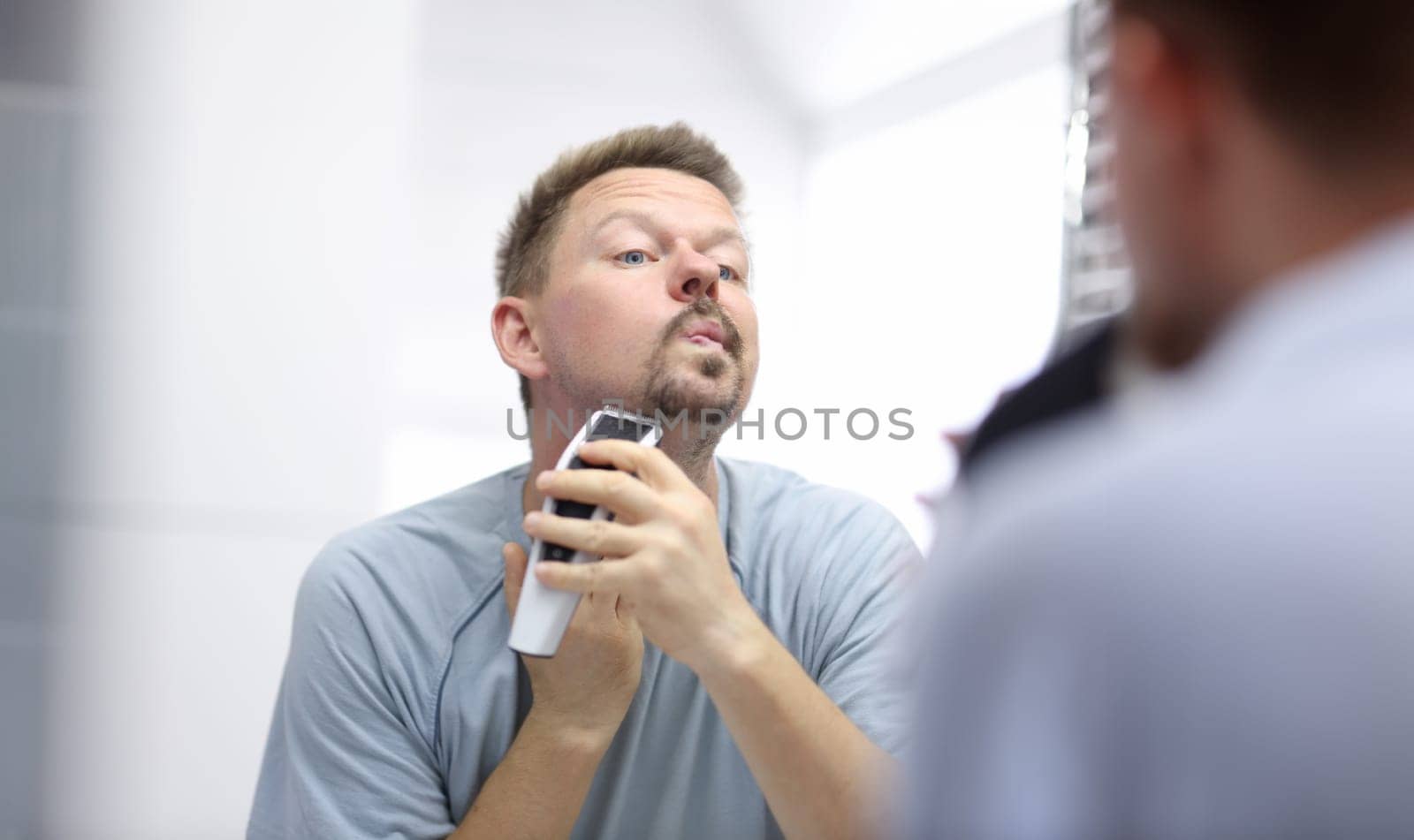 Young man shaves his face with razor while looking in mirror in bathroom. Daily morning hygiene procedures for men concept.