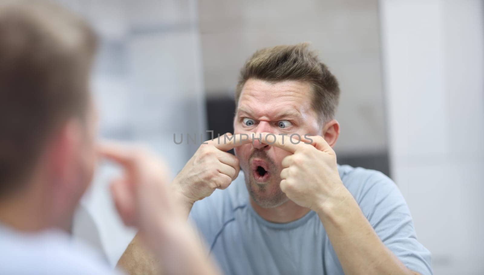 Young man looks in mirror and presses pimples on his nose. Facial skin care procedures at home concept.