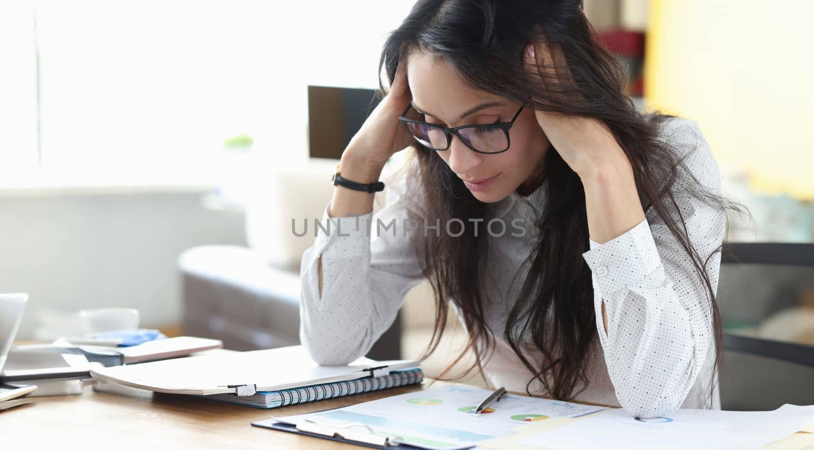 Tired woman with glasses sit at table and massages her head with her hands and think. Employee examines graphs and profit charts in office.