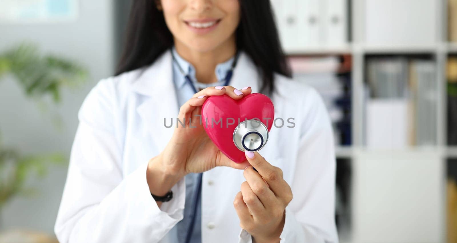 Hands of female GP holding stethoscope head near red toy heart as cardiac problems prophylaxis and recovery symbol closeup