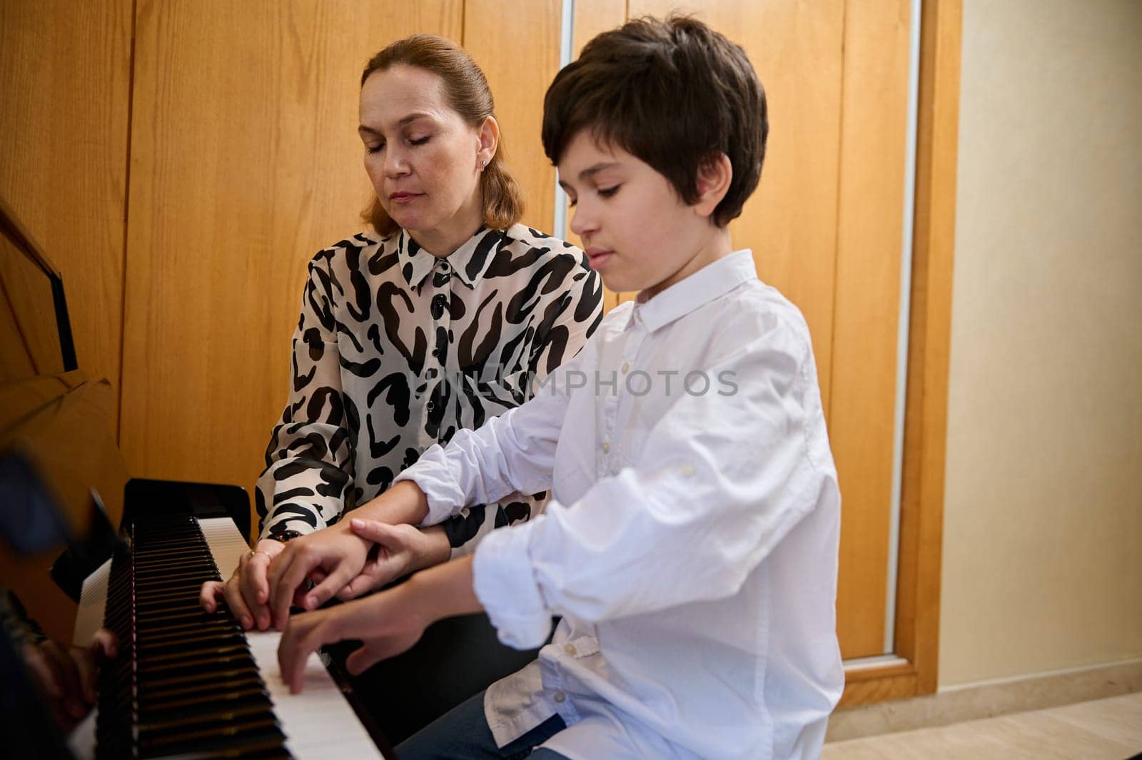 Handsome Hispanic boy in white casual shirt, learning playing piano, sitting at wooden piano forte and performing a musical composition under the guidance of is teacher. Musical education by artgf