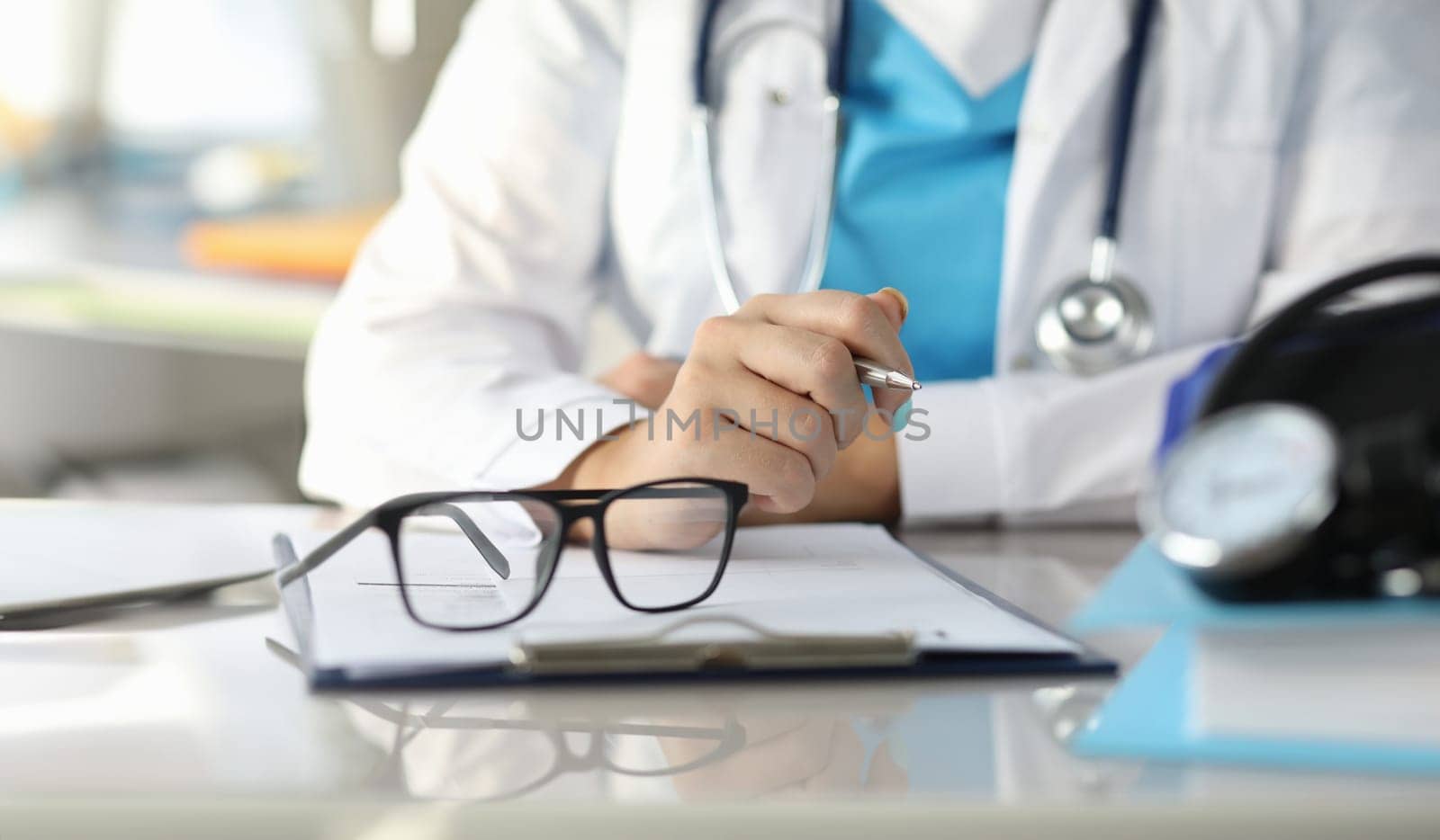 Close-up of glasses on working table. Clipboard with medical prescription and tonometer. Female doctor sitting in office at clinic. Special uniform. Modern medicine concept