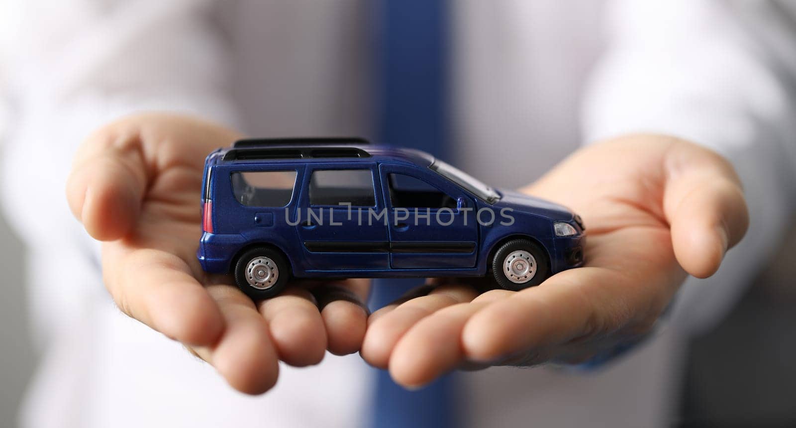 Closeup, man in shirt holds blue toy minivan car. Favorable car insurance. Inspection and diagnostics transport vehicles. International passenger transportation. Car sale from passenger compartment