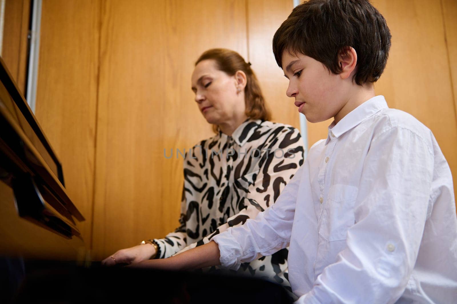 Confident teenage boy pianist musician creating the rhythm of melody, touching white and black keys of piano while playing pianoforte with his music teacher , sitting nearby on the blurred background by artgf