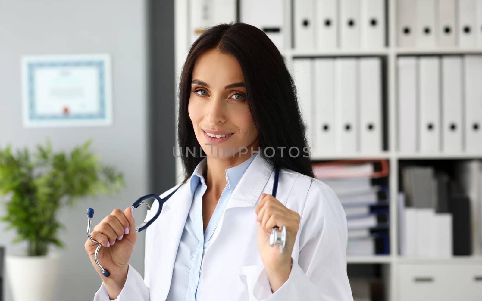 Pretty adult caucasian female GP stretching stethoscope in hands with pride facial expression looking in camera headshot