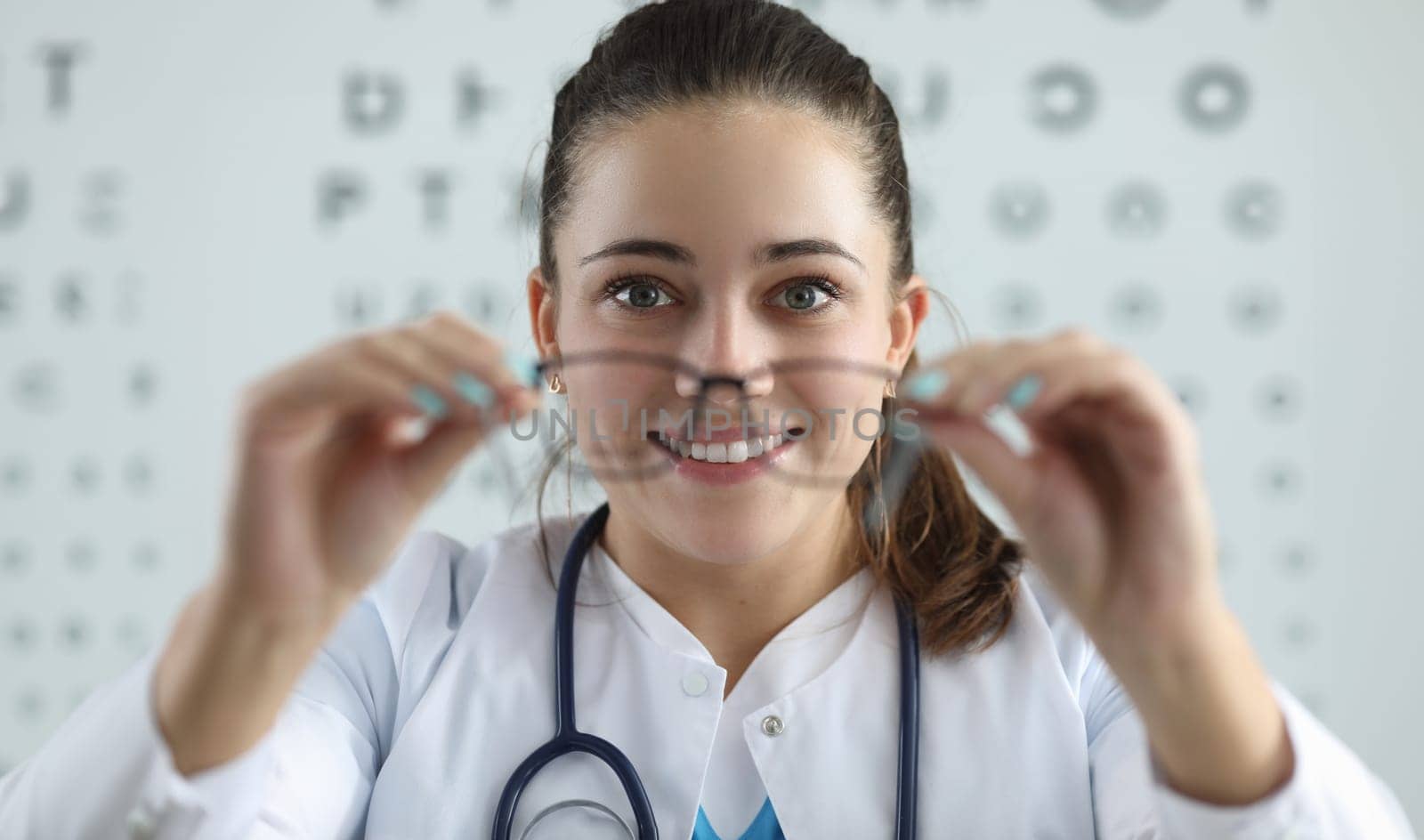 Portrait of cheerful female ophthalmologist try on vision glasses. Smiling young eye doctor check eyesight with special board. Modern medicine and healthcare concept
