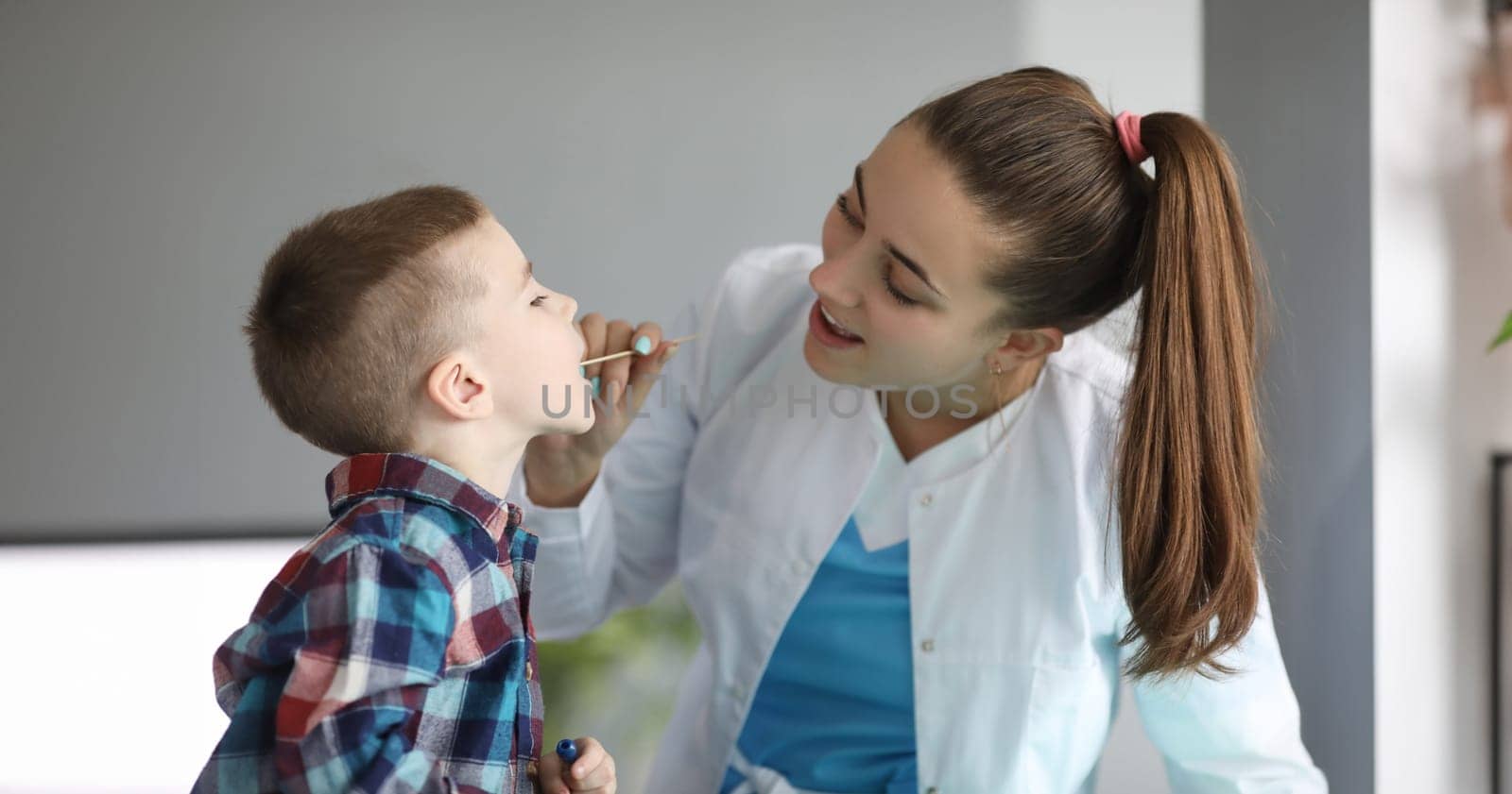 Close-up of lovely young doctor checking child throat at planned appointment. Entertaining kid and examine health. Professional pediatric and medicine concept