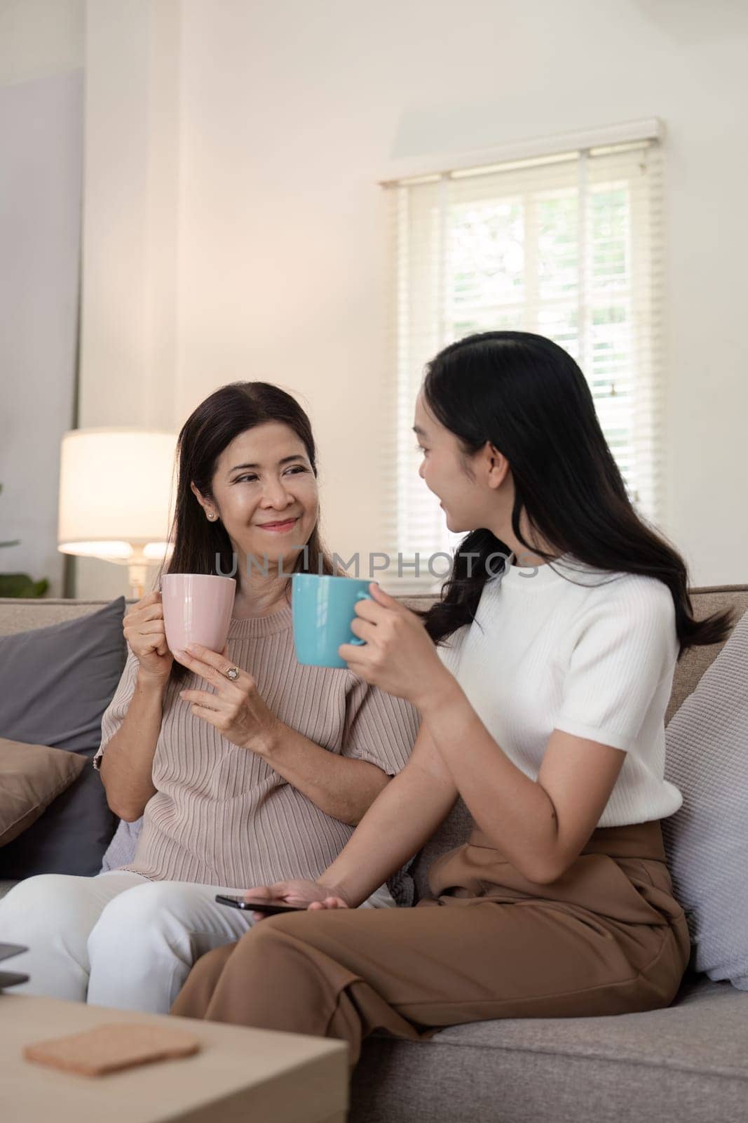 Happy senior mother with adult daughter sitting on couch and holding cups with coffee or tea at home. Enjoy family concept.