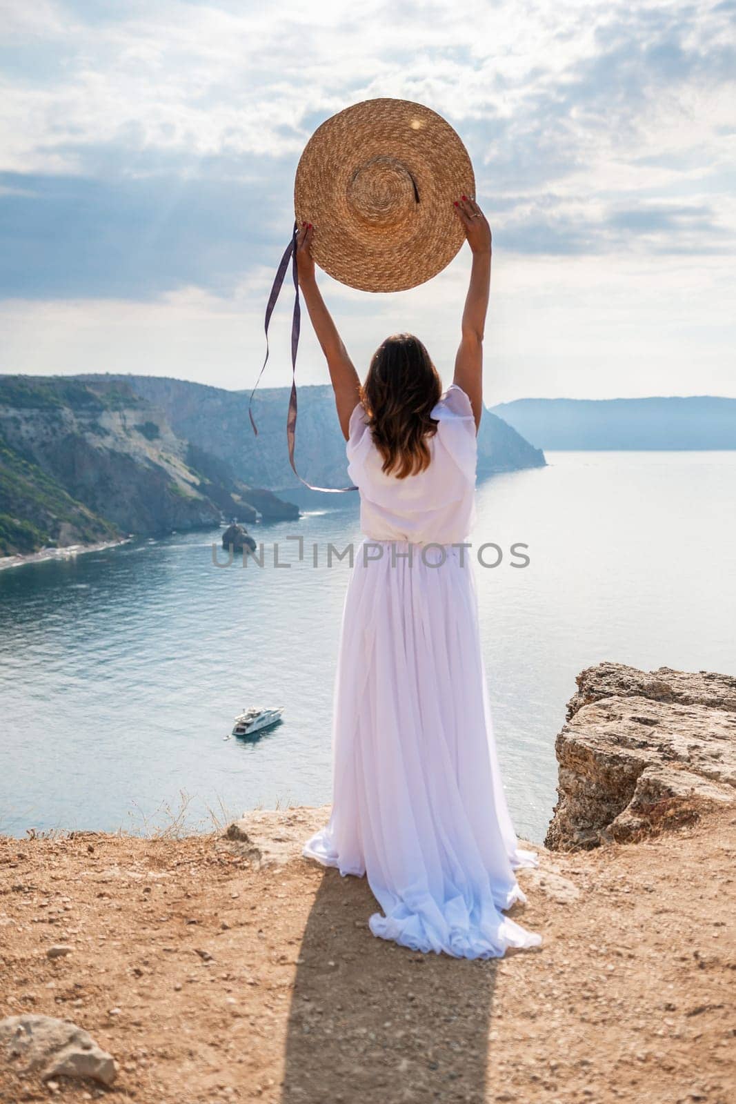 woman white dress stands on a rocky cliff overlooking the ocean. She is wearing a straw hat and she is enjoying the view