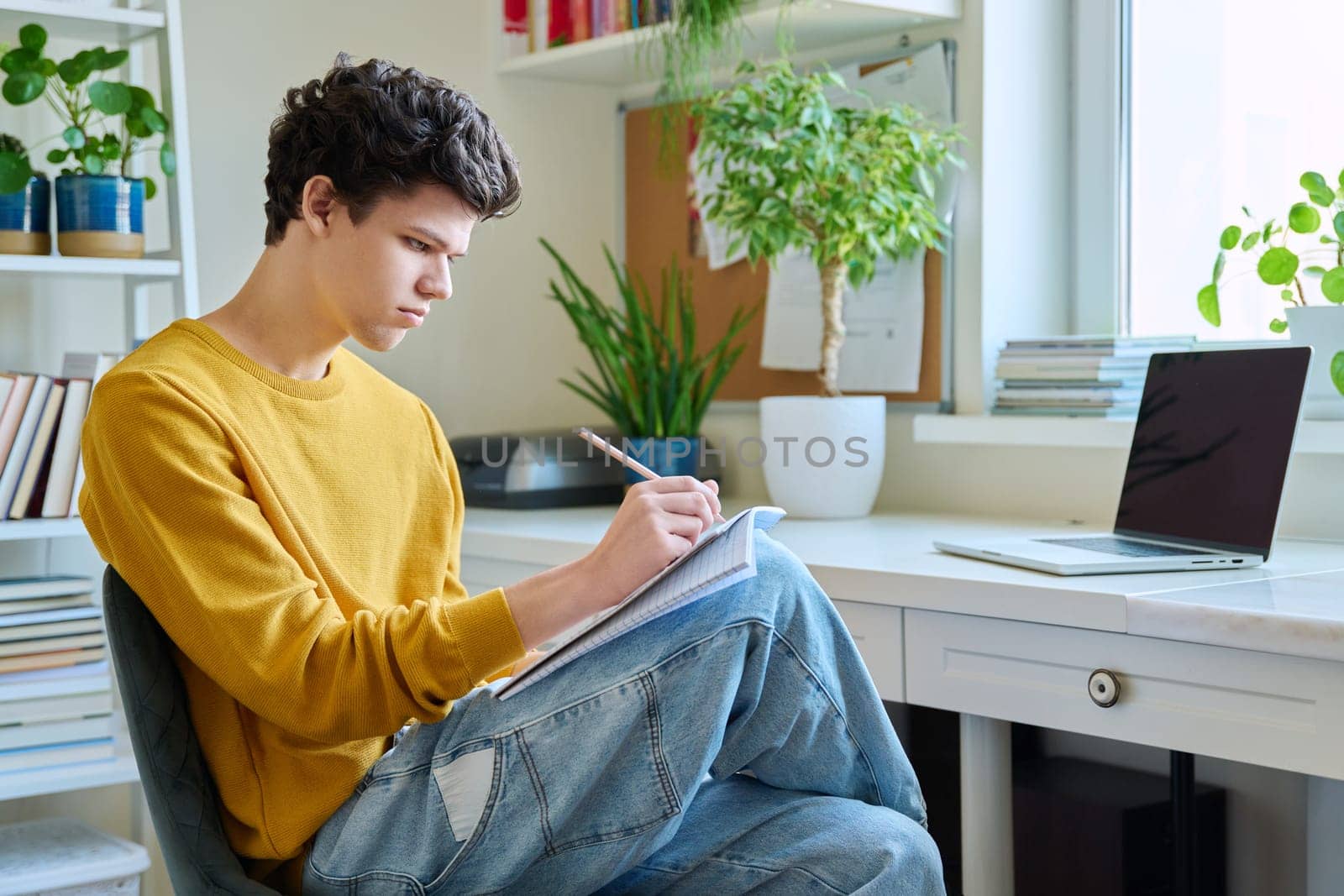 Guy college student sitting at home, looking at laptop screen, making notes in notebook by VH-studio