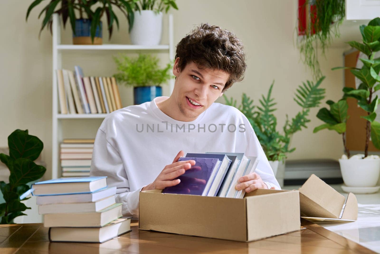 Satisfied young male customer buyer sitting at home unpacking cardboard box parcel with new books, online purchases. Delivery by mail, internet store bookstore