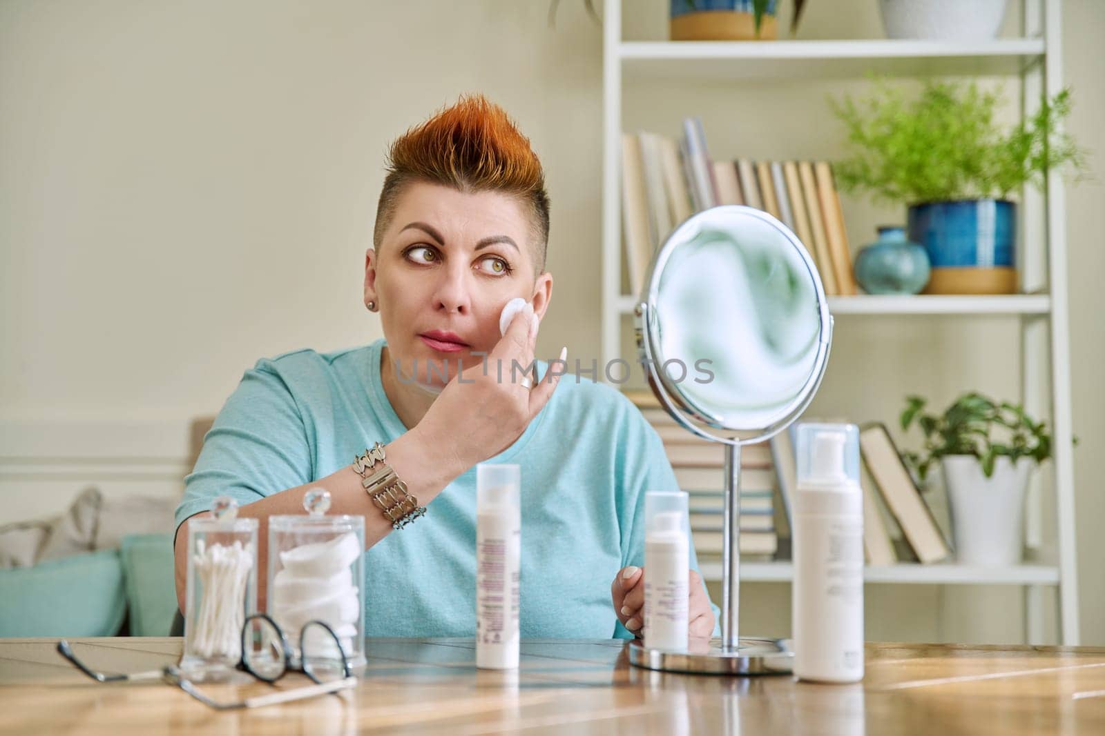 Middle-aged woman with a mirror taking care of her facial skin by VH-studio