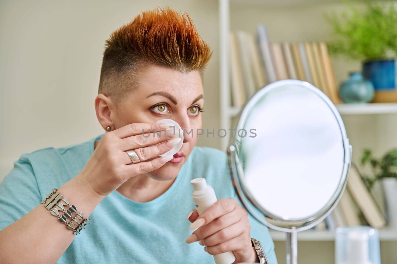 Middle-aged woman with a mirror taking care of her facial skin by VH-studio