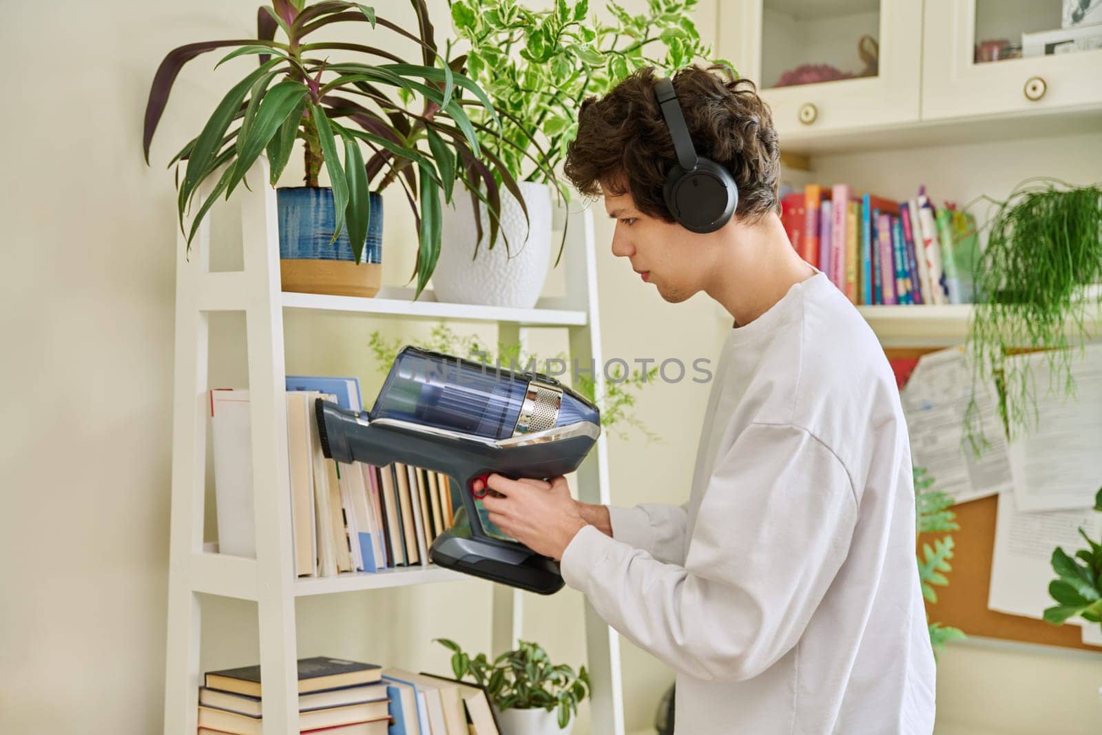 Young man wearing headphones vacuums bookshelves at home by VH-studio