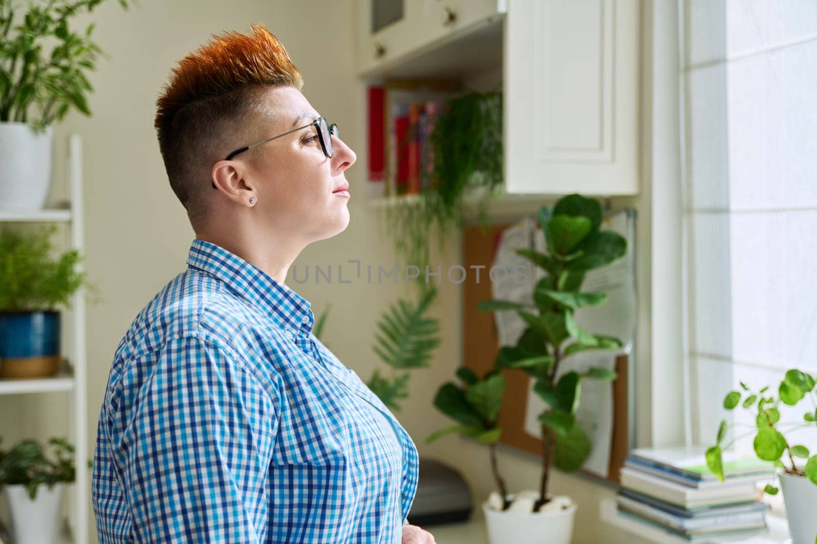 Profile portrait of serious middle-aged woman in glasses with red haircut looking at window in home interior, copy space. Mature people, lifestyle, health, life concept