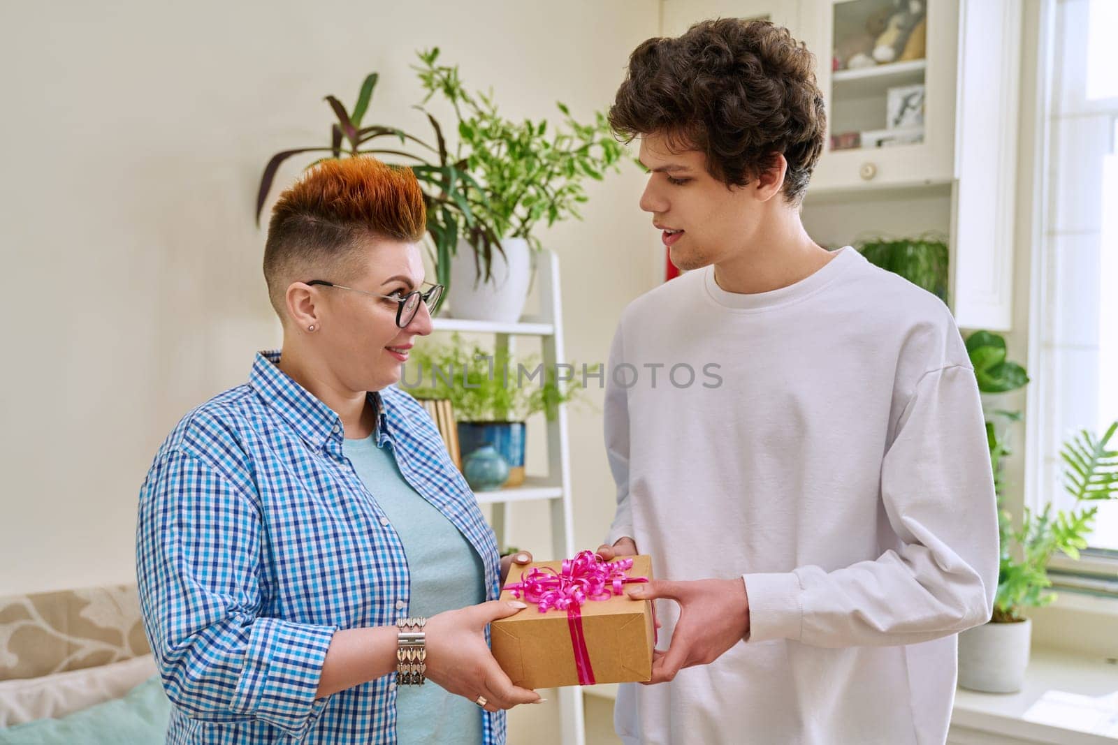 Cheerful teenage son congratulating middle aged mother with surprise gift, standing together at home. Holiday, birthday, congratulations, love, mother's day, happiness, joy, family concept