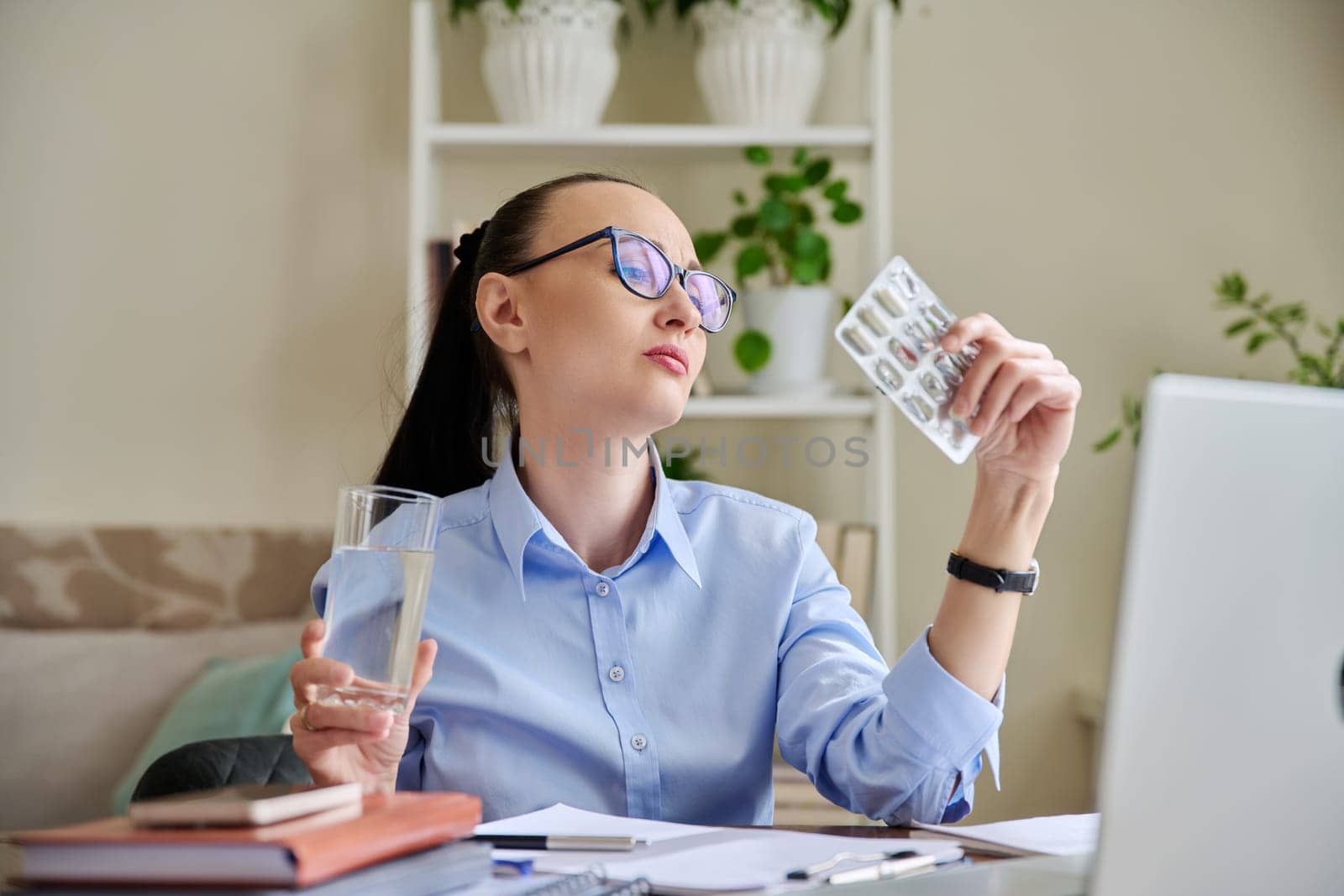 Serious woman at workplace holding blister with capsules glass of water. Female taking painkillers vitamins nutritional supplements antiviral drugs antioxidants antidepressants. Pharmacy health care