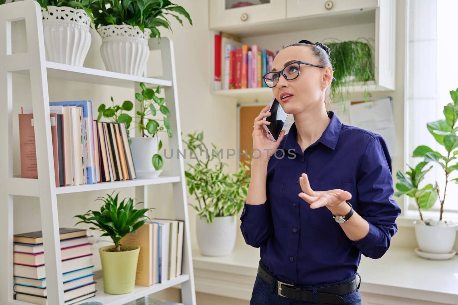 Beautiful emotional woman in her 30s talking on a cell phone, standing in her room at home by VH-studio