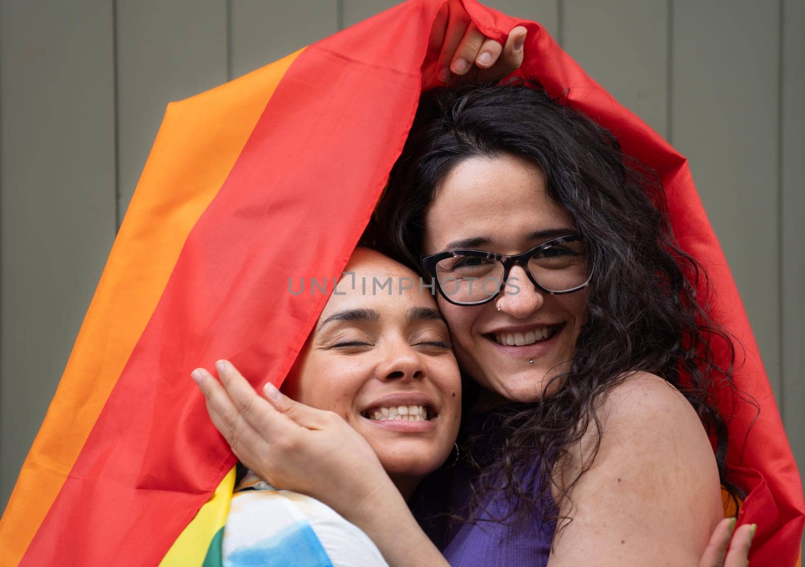 Lesbian couple are hugging and caressing under a rainbow flag with happy expression.