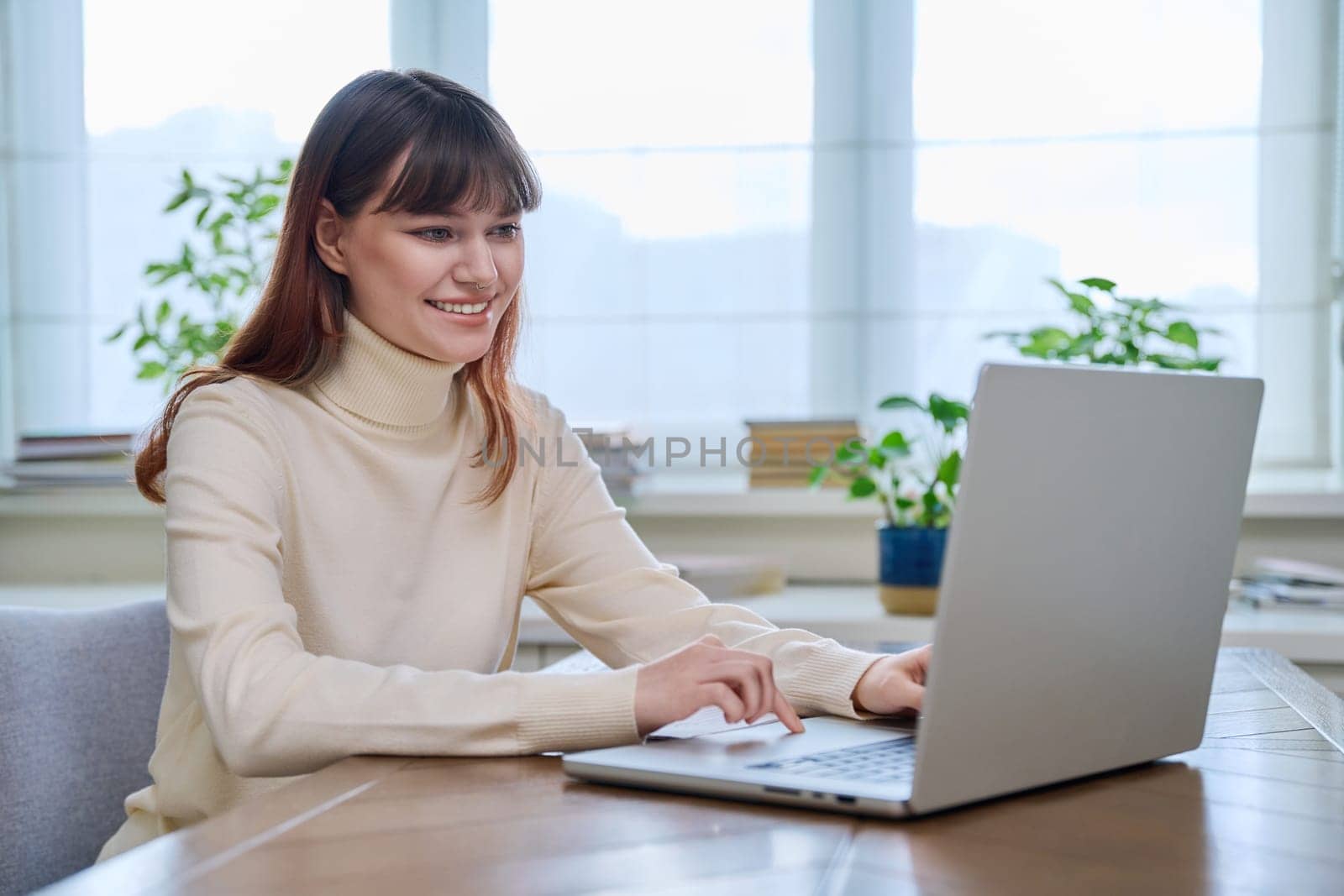 College student girl sitting at desk using laptop computer, typing on a keyboard, at home. Teenager female watching webinar, preparing for exam tests, studying remotely.