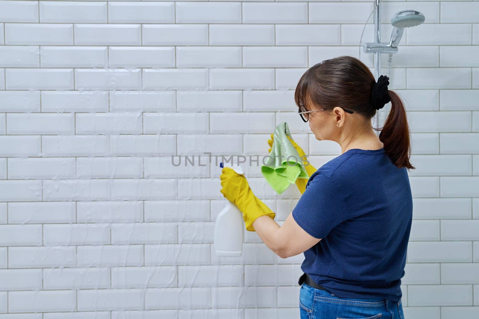 Woman with detergent spray, washcloth cleaning in bathroom, washing white wall tiles. Routine house cleaning, home hygiene, housecleaning service, housekeeping, housework concept