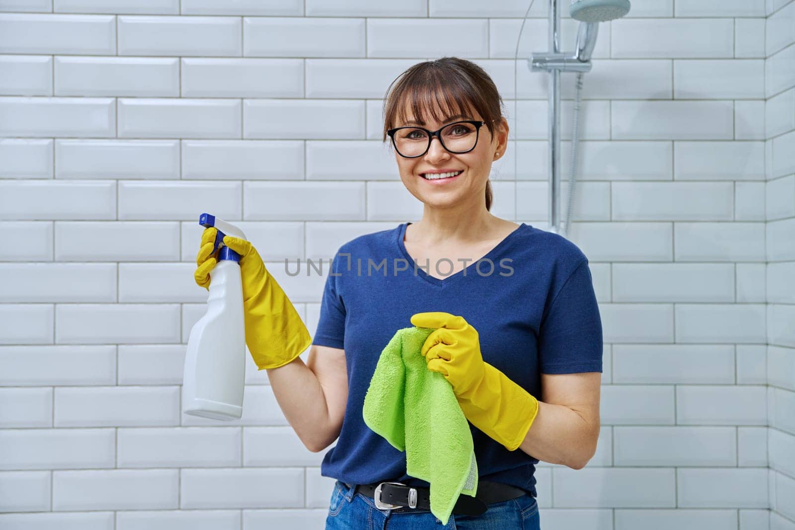 Middle-aged woman in gloves with detergent spray washcloth doing cleaning in bathroom, smiling looking at camera. Routine house cleaning, home hygiene, housecleaning service, housekeeping, housework