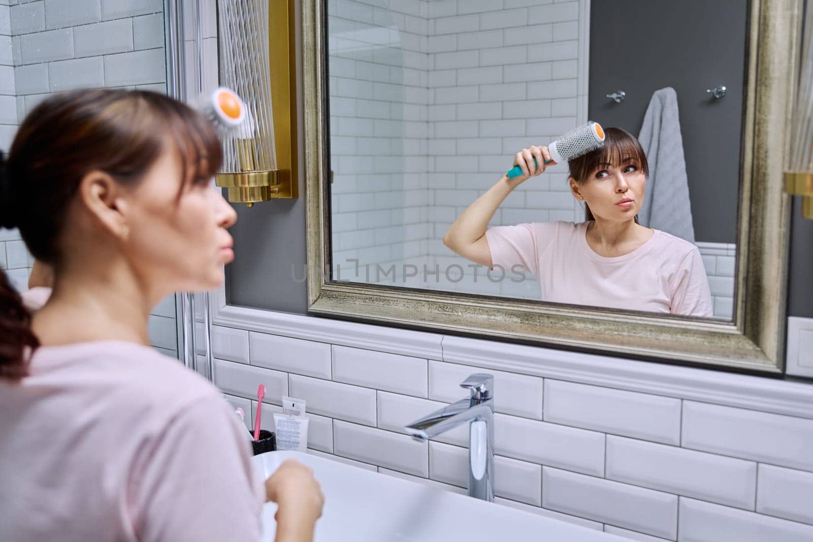 Middle-aged woman with hairspray comb looking in mirror, doing hair styling, in bathroom. Beauty, age, cosmetics, hair accessories