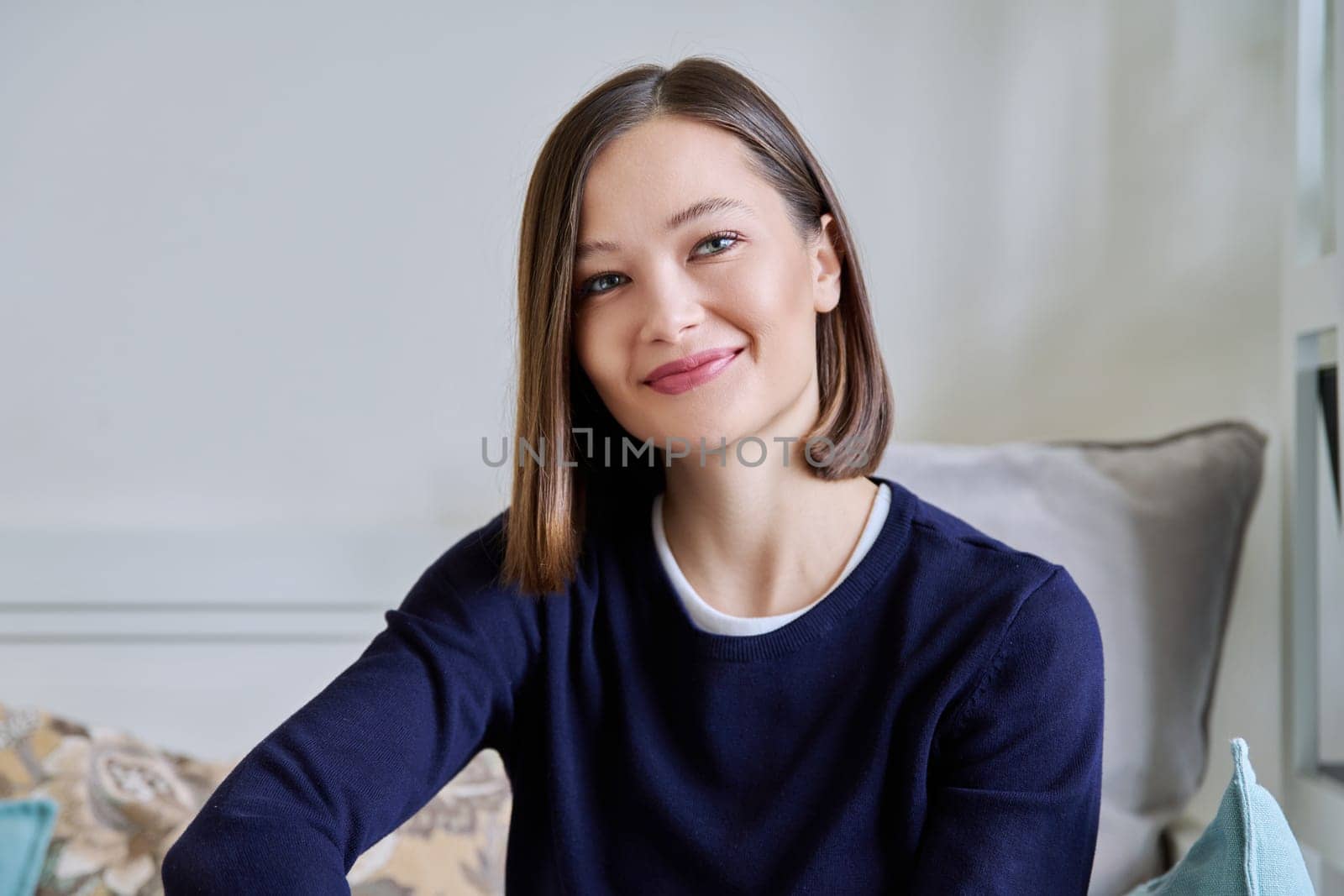 Portrait of young attractive smiling woman in home interior. Happy cheerful relaxed 20s female looking at camera, sitting on couch. Beauty, youth, happiness, health, lifestyle concept