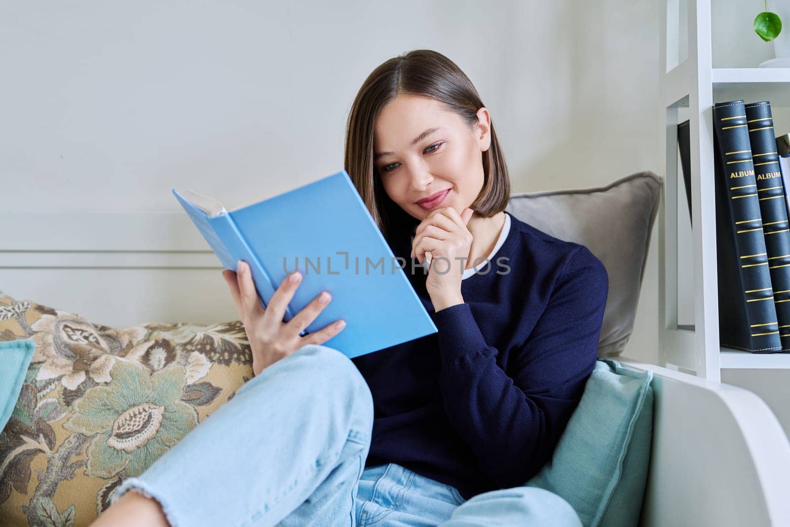 Young woman reading paper book, relaxed sitting on sofa at home. Fiction, hobby, leisure, youth concept