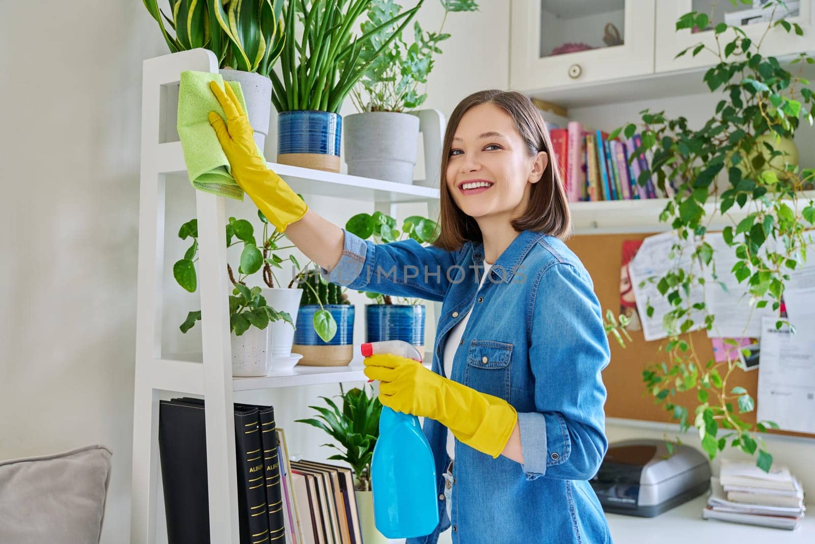 Young woman doing house cleaning, wiping dust, wearing gloves with cleaning detergent agent spray rag in room interior. Routine home cleaning, cleanliness housework housekeeping household lifestyle