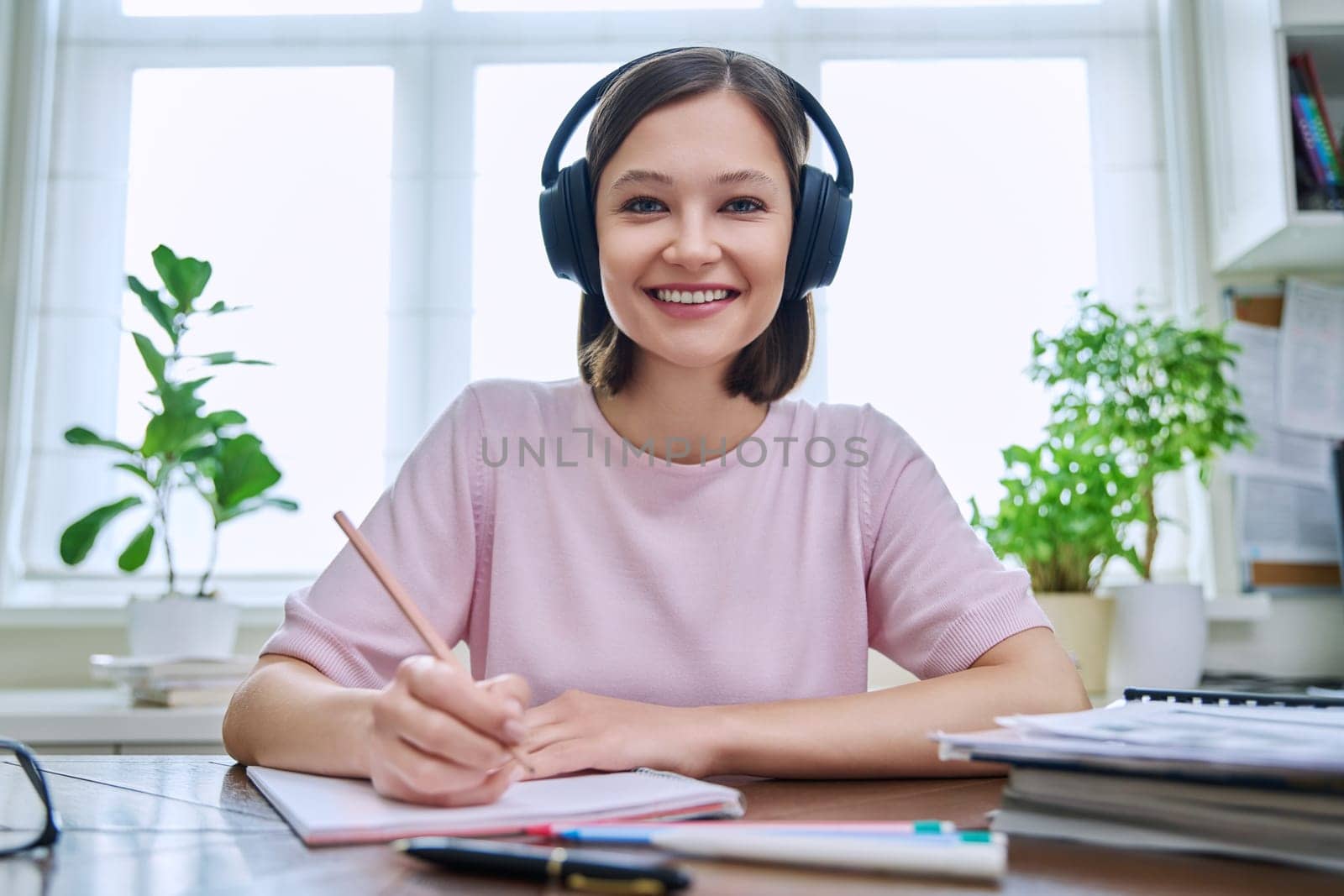 Webcam portrait of young 20s female student wearing headphones, sitting at home at desk, looking at camera, listening. Internet online webinar, video chat call conference, technology education training