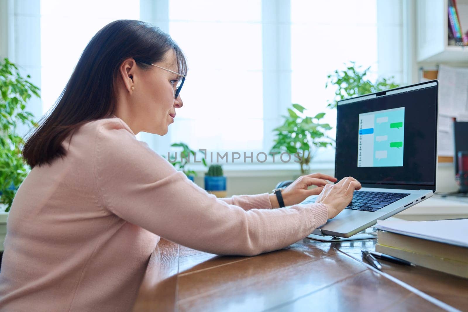 Middle-aged woman communicates online using laptop at her desk at home. Chat on computer screen, communication work leisure technology concept