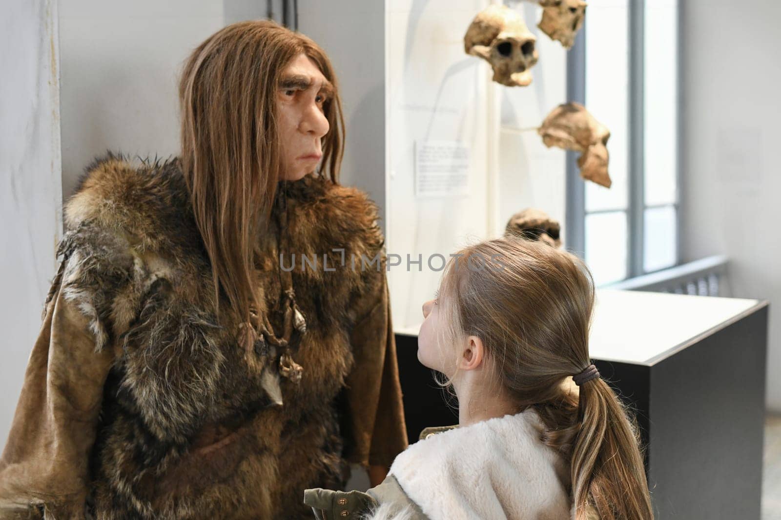 The girl looks at wax figure of a Neanderthal with long hair in skins by Godi