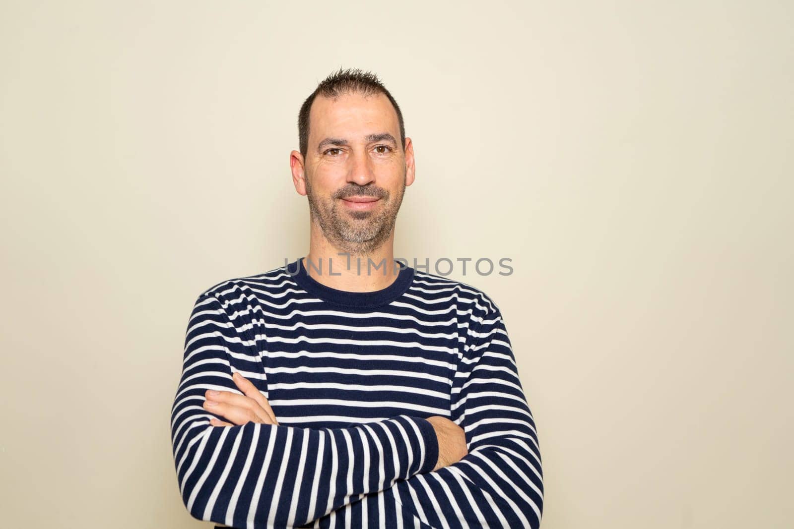 Bearded Hispanic man in his 40s wearing a striped sweater keeping his arms crossed and smiling while isolated on beige background. by Barriolo82