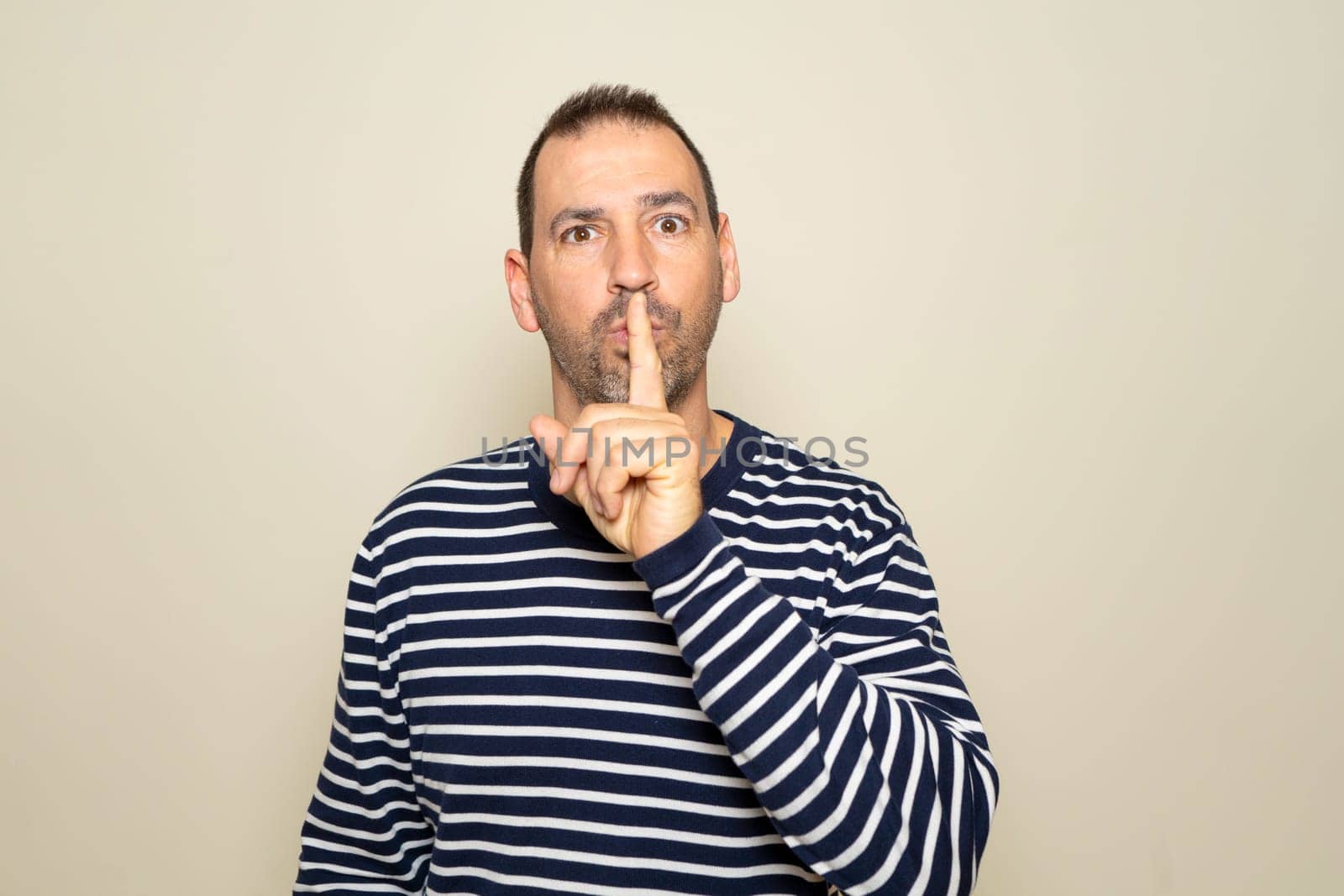 Hispanic man with a beard in his 40s with a striped sweater putting his index finger to his mouth asking for silence, he is keeping a secret. Isolated on beige studio background