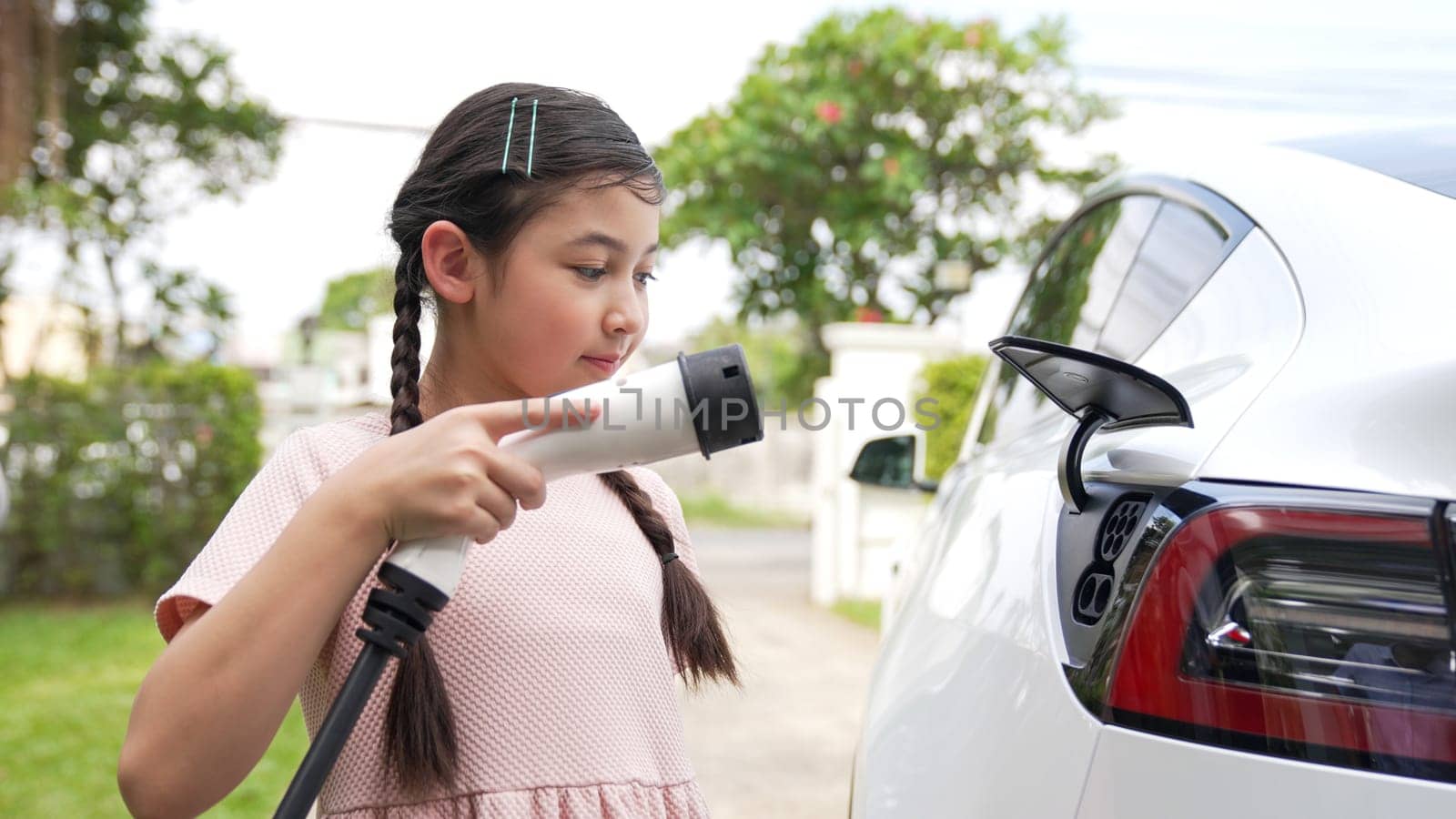 Happy little young girl learn about eco-friendly and energy sustainability as she recharge electric vehicle from home EV charging station. EV car and sustainable future generation concept. Synchronos