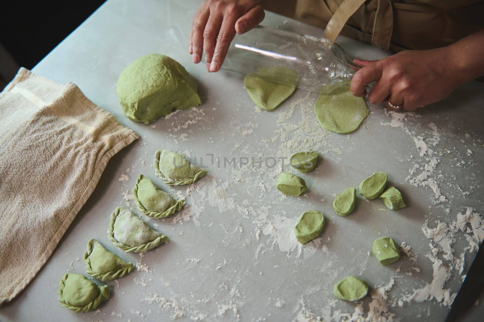 Top view female housewife's hands using a glass wine bottle for rolling out the dough with green spinach, cooking traditional Ukrainian Varennyky with mashed potatoes. Food. Traditions. Culture