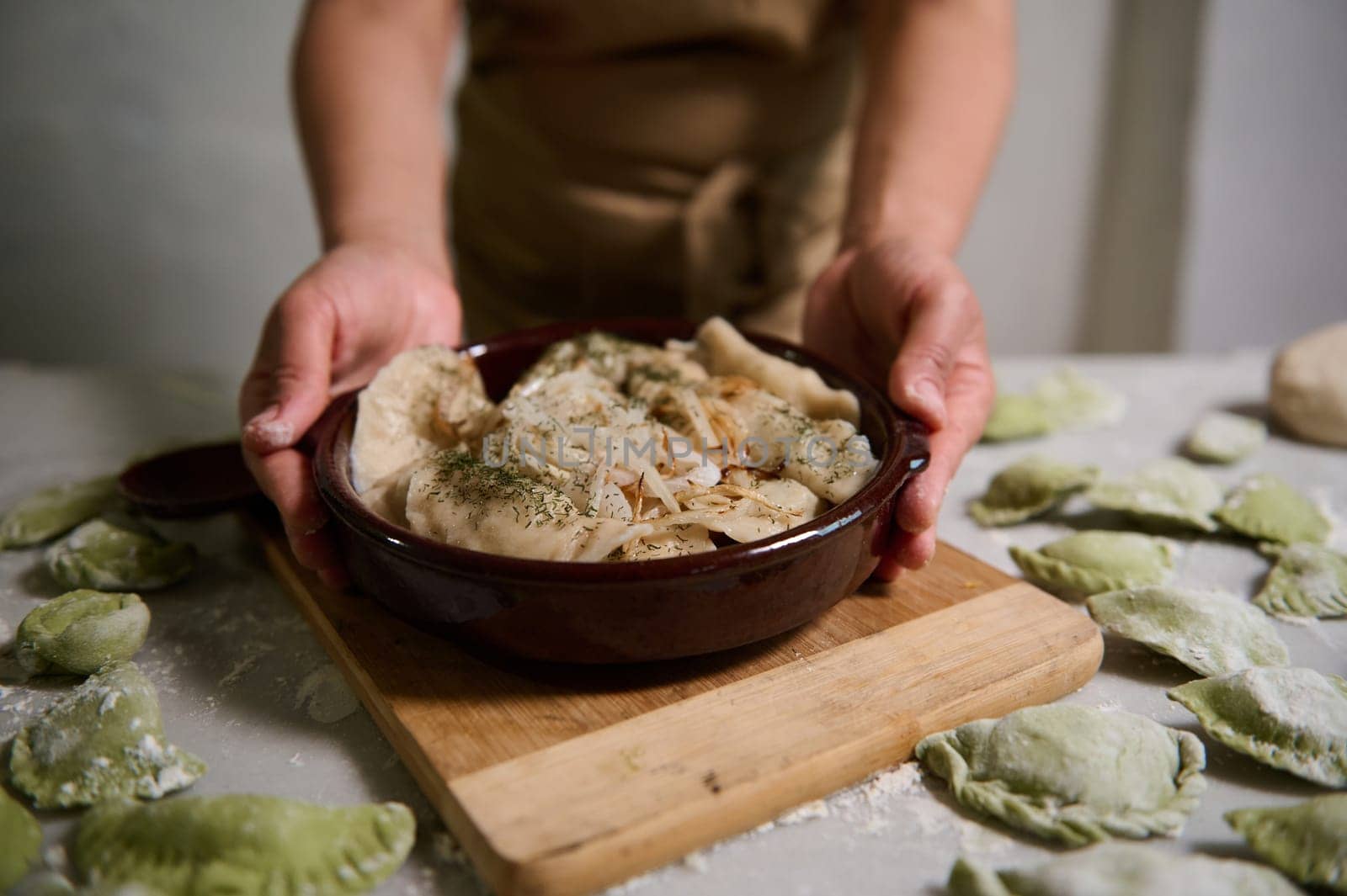 Close-up of the hands of housewife serving delicious homemade dumplings in a clay dish, standing at floured marble kitchen table with raw uncooked varennyki. Homemade food and ethnic culinary concept