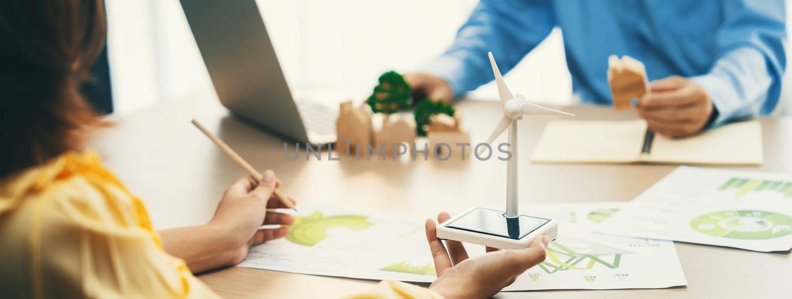 Cropped image of businesswoman presents eco-friendly house by using green design to manager while holding the wooden house block at table with windmill model placed with document. Delineation.