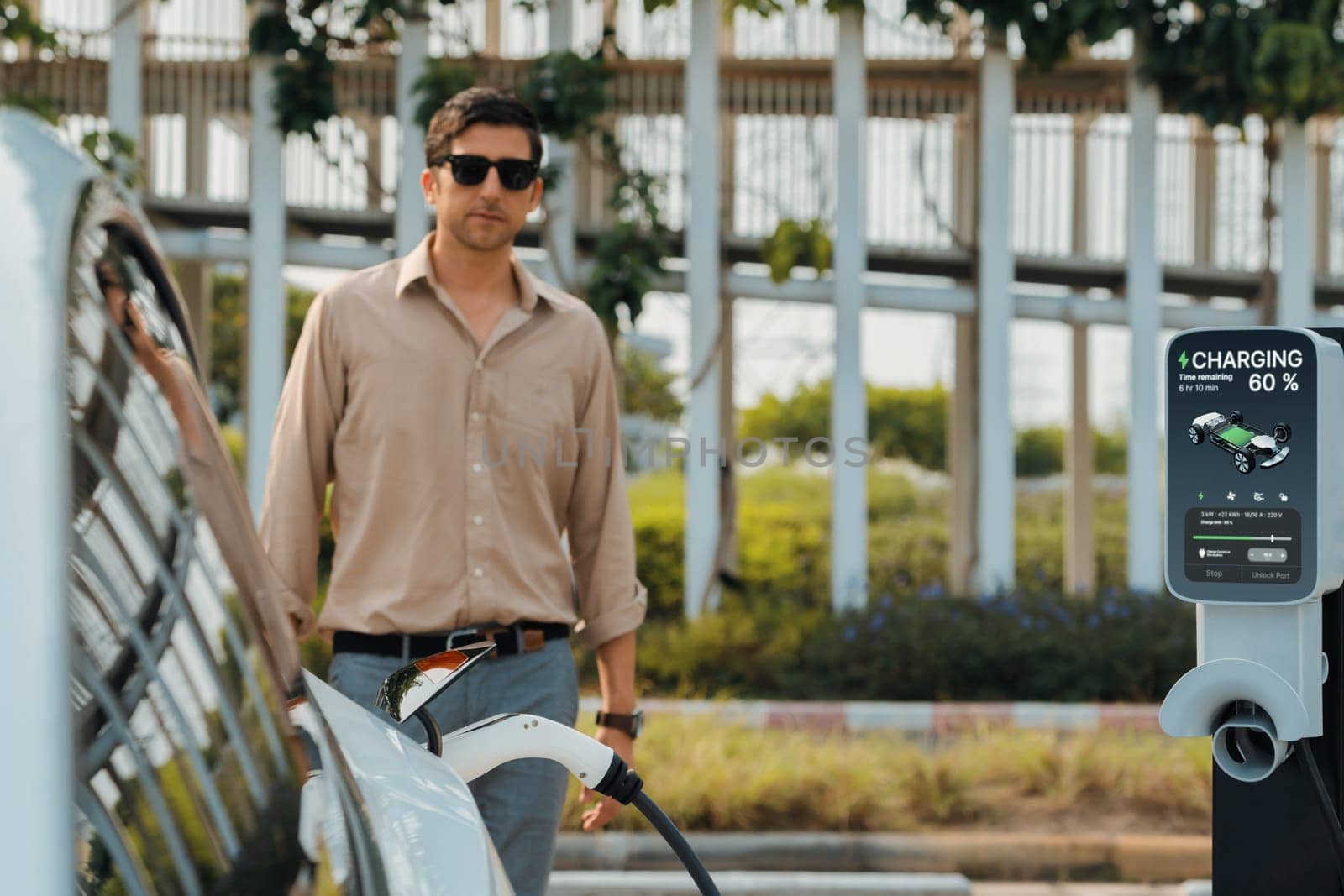 Young man put EV charger to recharge electric car's battery from charging station in city commercial parking lot. Rechargeable EV car for sustainable environmental friendly urban travel. Expedient