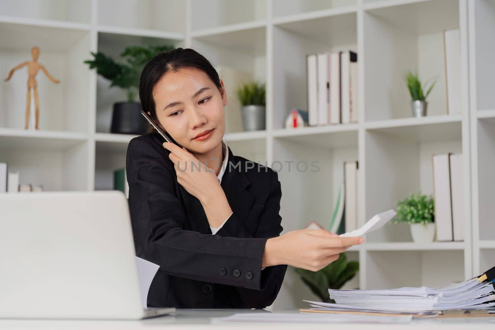 Business woman looking busy while talking on mobile and searching through document on messy table in an office.