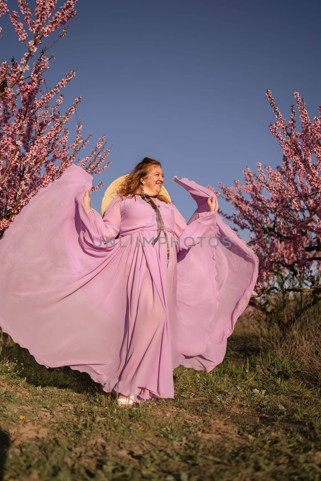 Woman blooming peach orchard. Against the backdrop of a picturesque peach orchard, a woman in a long pink dress and hat enjoys a peaceful walk in the park, surrounded by the beauty of nature. by Matiunina