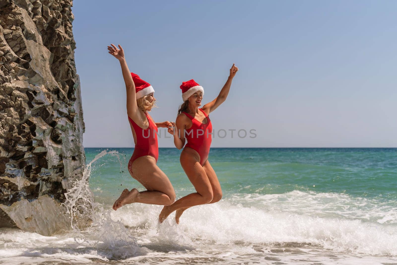 Women Santa hats ocean play. Seaside, beach daytime, enjoying beach fun. Two women in red swimsuits and Santa hats are enjoying themselves in the ocean waves and raising their hands up